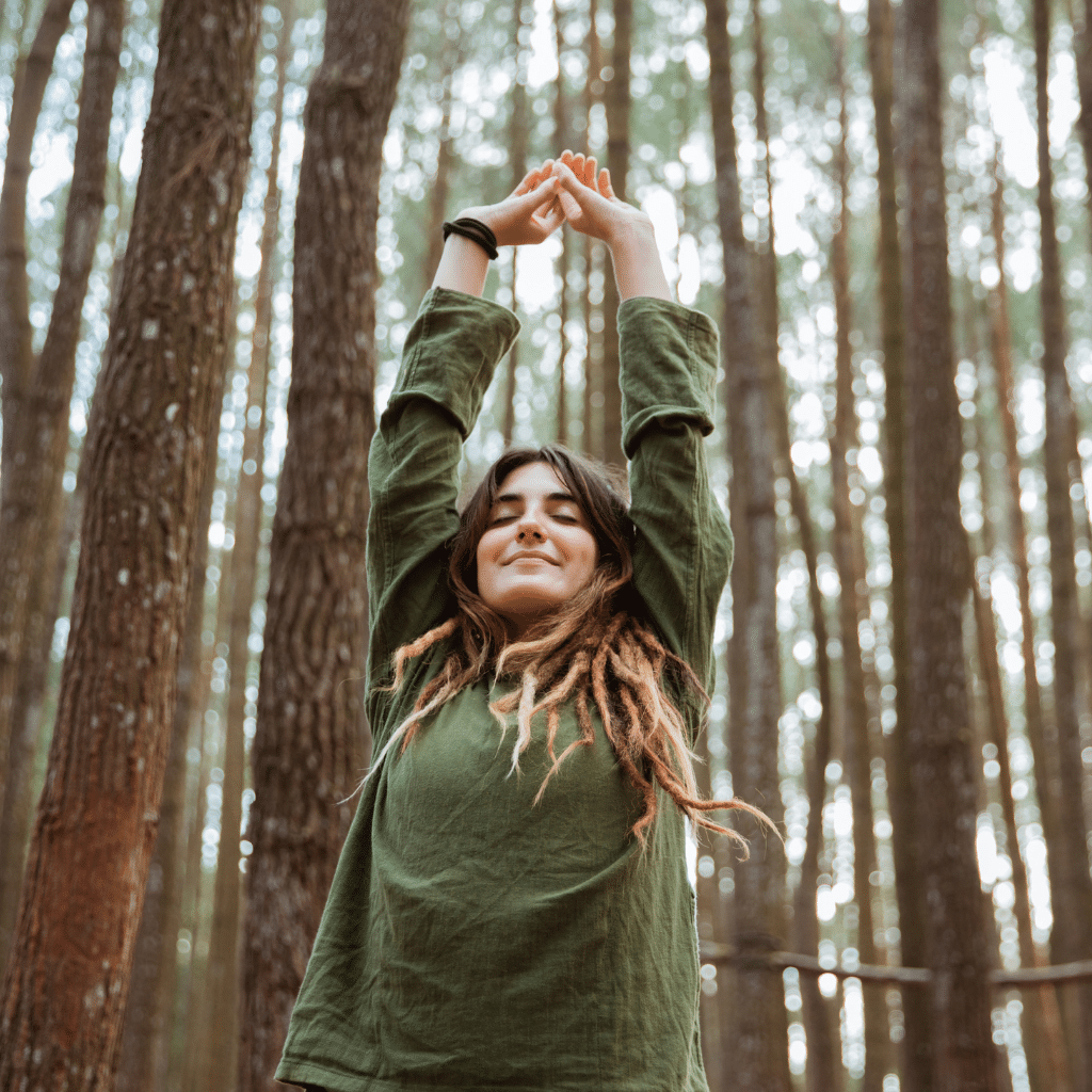 Woman wearing olive green clothing stretching with hands above her head in a forest.